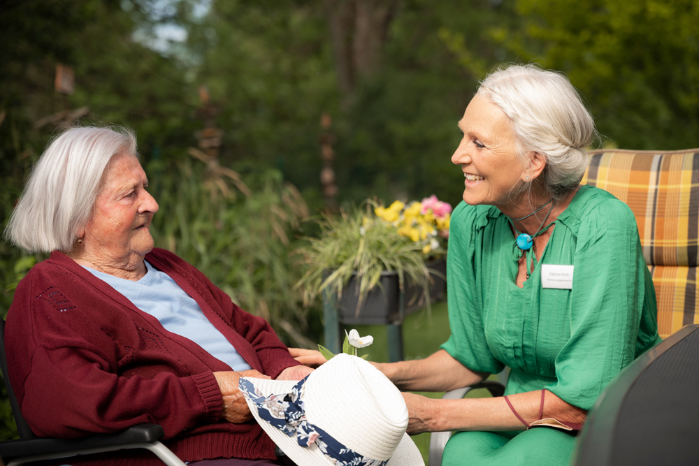 Eine lächelnde Frau (Ehrenamtliche oder Betreuungskraft) unterhält sich liebevoll mit einer Seniorin im Garten, symbolisiert die wertvolle Betreuung, das soziale Engagement und die menschliche Begleitung bei Allershausen Care.